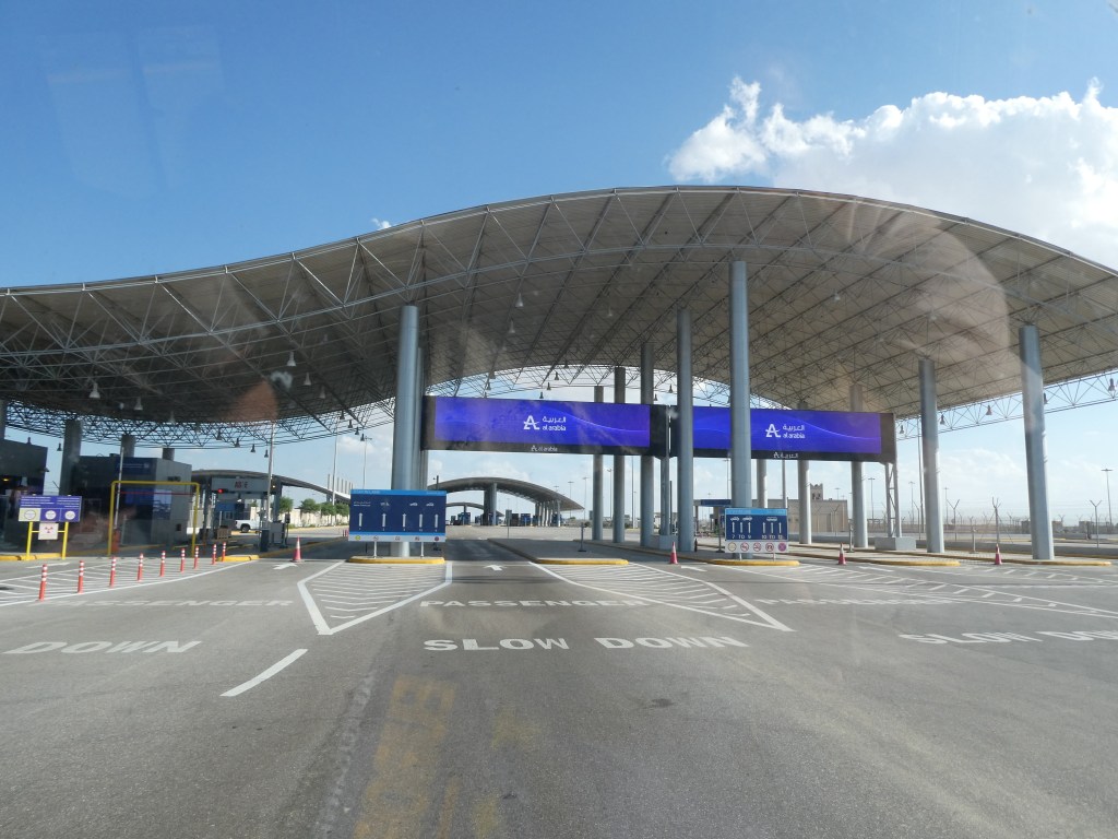 Contrôle frontalier à l'entrée de l'Arabie Saoudite, avec un bâtiment moderne et des panneaux de signalisation indiquant de ralentir.