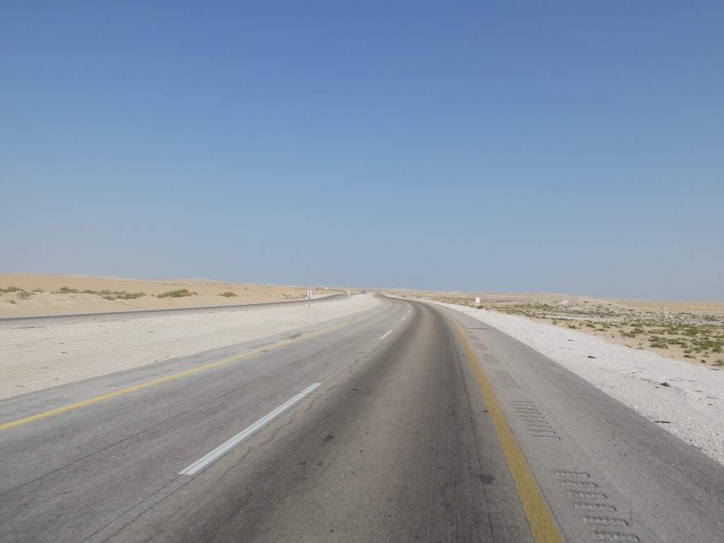 Une route désertique bordée de sable et de petites végétations sous un ciel bleu clair.