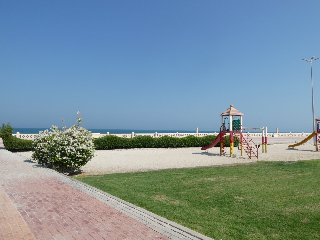 Un parc de jeux avec toboggan et balançoires, entouré de verdure, offrant une vue sur la mer sous un ciel bleu dégagé.