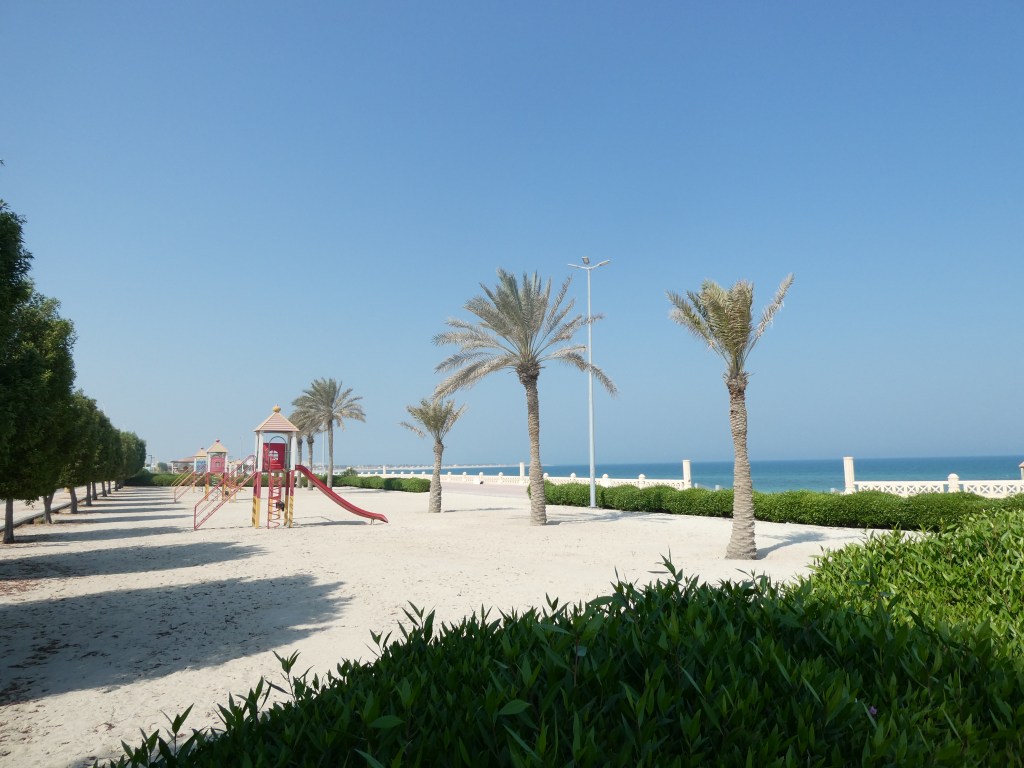 Une vue d'un parc de plage avec des palmiers, une structure de jeux pour enfants et la mer en arrière-plan dans un ciel ensoleillé.