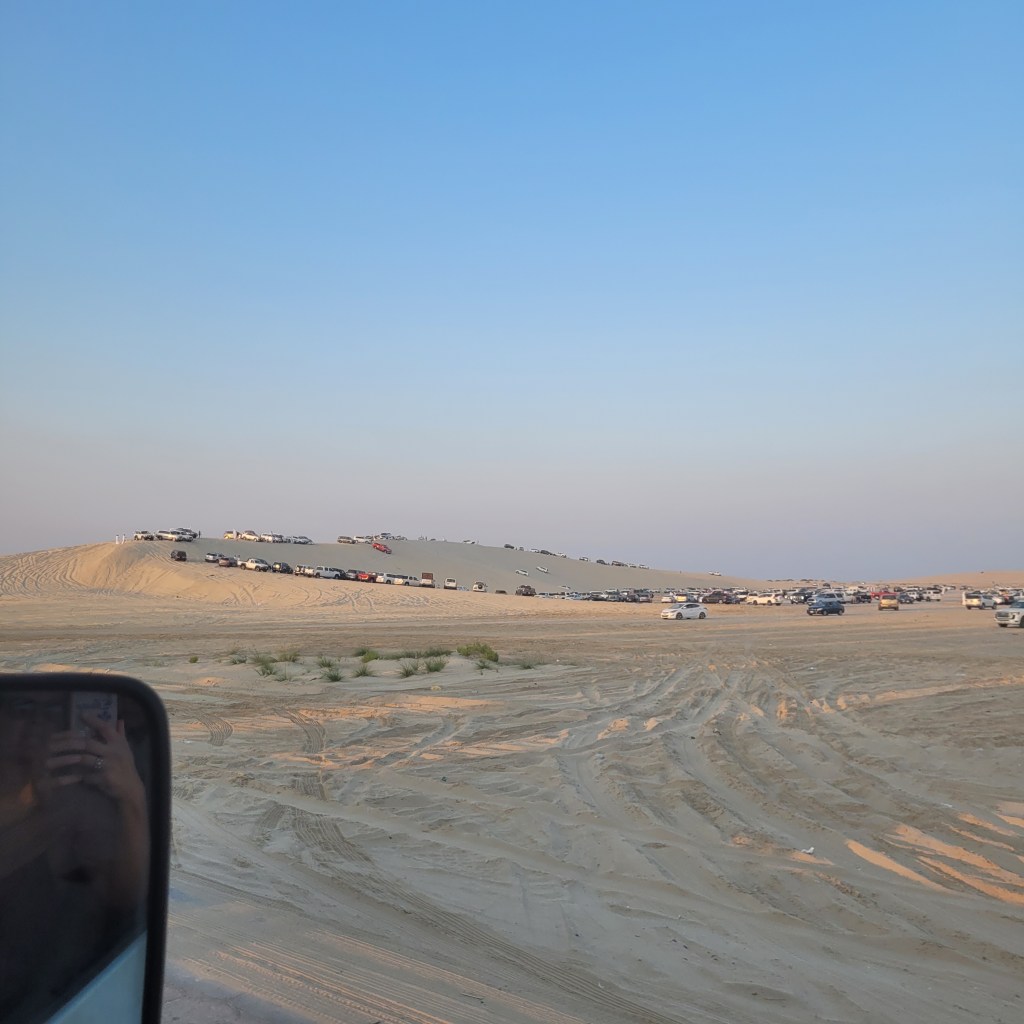 Un vaste paysage désertique avec une dune de sable surmontée de nombreux véhicules garés, un ciel bleu clair en arrière-plan.