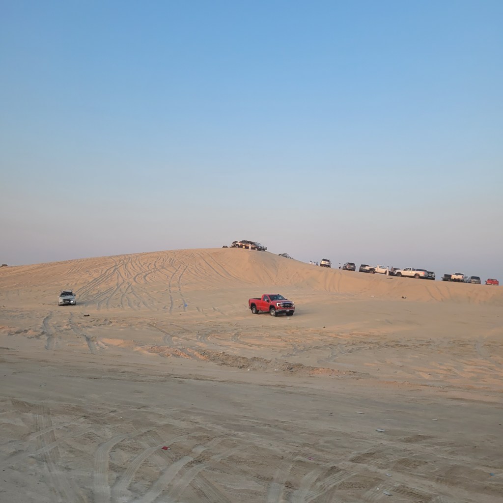 Vue d'un paysage désertique avec plusieurs véhicules stationnés sur une dune de sable au coucher du soleil.