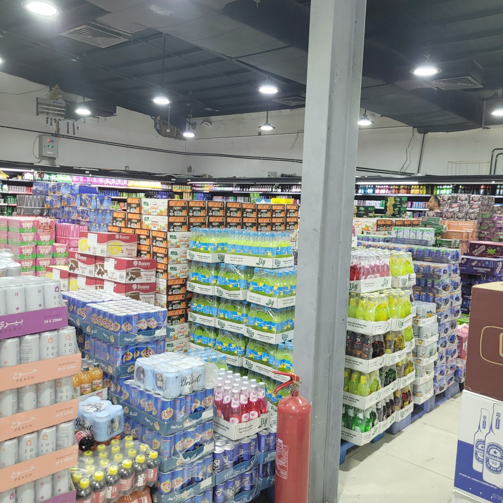 Intérieur d'un supermarché en Arabie Saoudite, avec des étagères remplies de boissons et de produits alimentaires variés, et un extincteur visible.