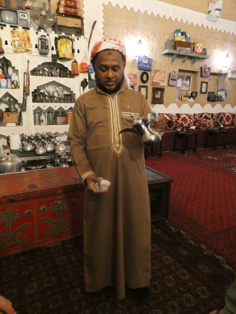 Un homme en tenue traditionnelle saoudienne présente une théière en métal, entouré d'une décoration intérieure orientale.