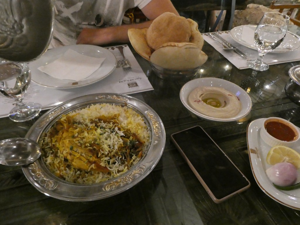 Un plat traditionnel saoudien présentant du riz jaune, viande, et divers accompagnements, servi dans un plateau décoratif en métal. À côté, des pains, un petit bol de hummus et une assiette avec des sauces et légumes, le tout sur une table bien dressée.