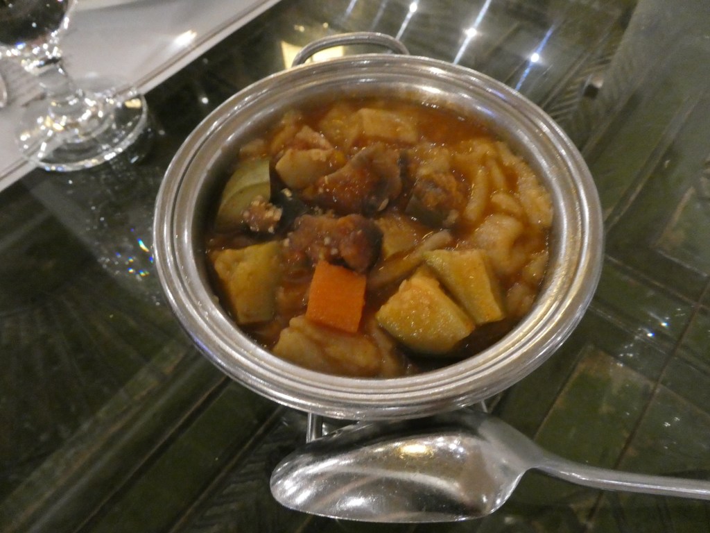 Un plat traditionnel saoudien de ragoût de viande et de légumes servi dans une petite casserole en métal, accompagné d'une cuillère en métal, sur une table aux reflets brillants.