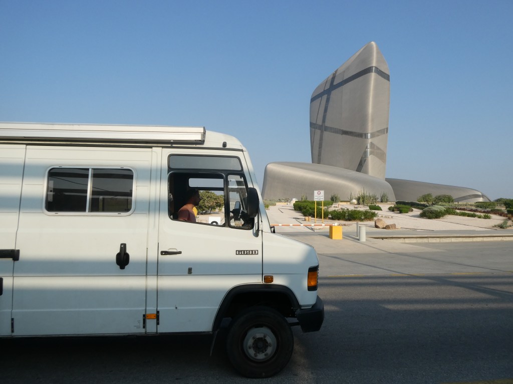 Une camionnette blanche garée près d'un bâtiment moderne aux formes architecturales dynamiques en Arabie Saoudite, le ciel est dégagé.