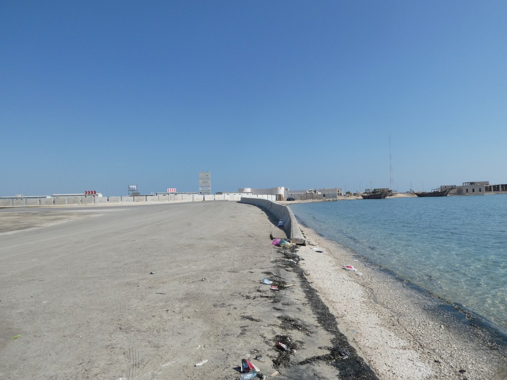 Vue d'une route côtière avec des déchets sur le sable et une mer claire à proximité sous un ciel bleu.