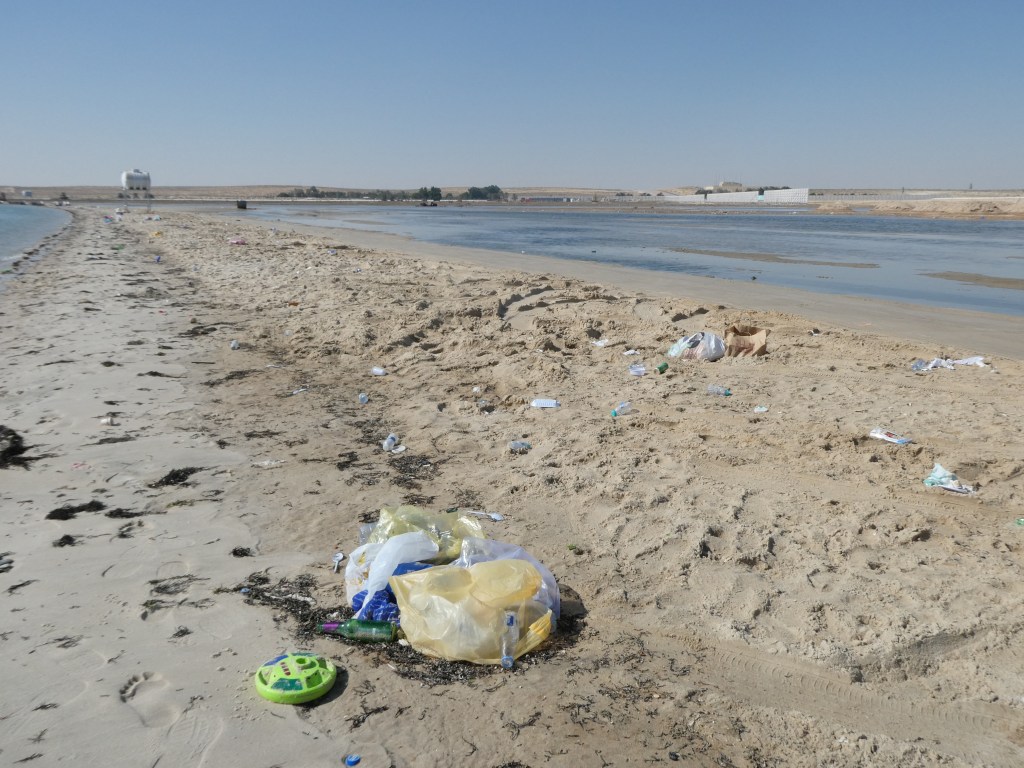 Plage en Arabie Saoudite jonchée de déchets, avec des sacs plastiques et divers détritus sur le sable.