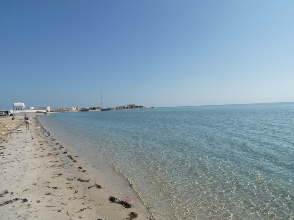 Vue de la plage avec des eaux calmes et un ciel bleu, un chemin de sable et des bâtiments en arrière-plan.