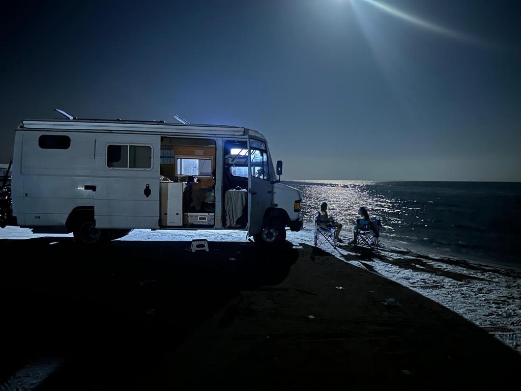 Un camping-car blanc stationné sur une plage la nuit, avec deux personnes assises sur des chaises près de l'eau, éclairé par la lune.