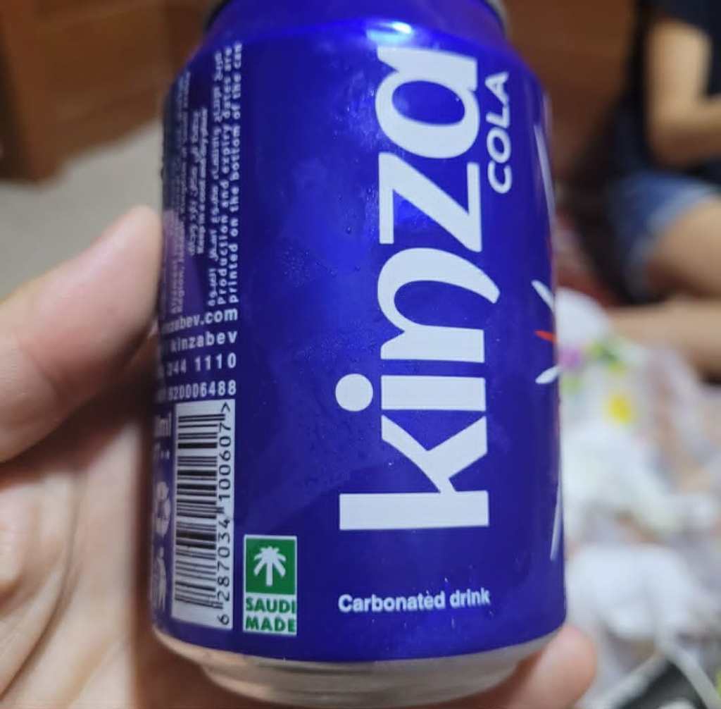 Une canette de Kinza Cola bleue, contenant une boisson gazeuse, avec des informations sur le produit et 'Fabriqué en Arabie Saoudite' visible.