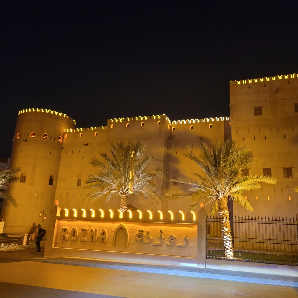 Vue de la forteresse Rozna illuminée la nuit, avec des palmiers en avant-plan et un panneau en bois gravé du nom 'Rozna'.