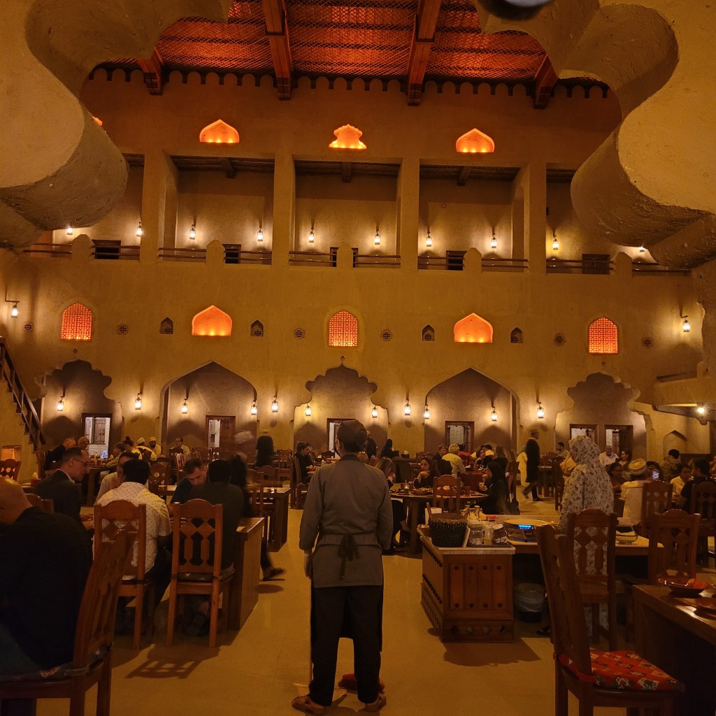 Intérieur d'un restaurant élégant avec des murs en terre, des lanternes et des fenêtres de style arabe, rempli de clients assis à des tables.