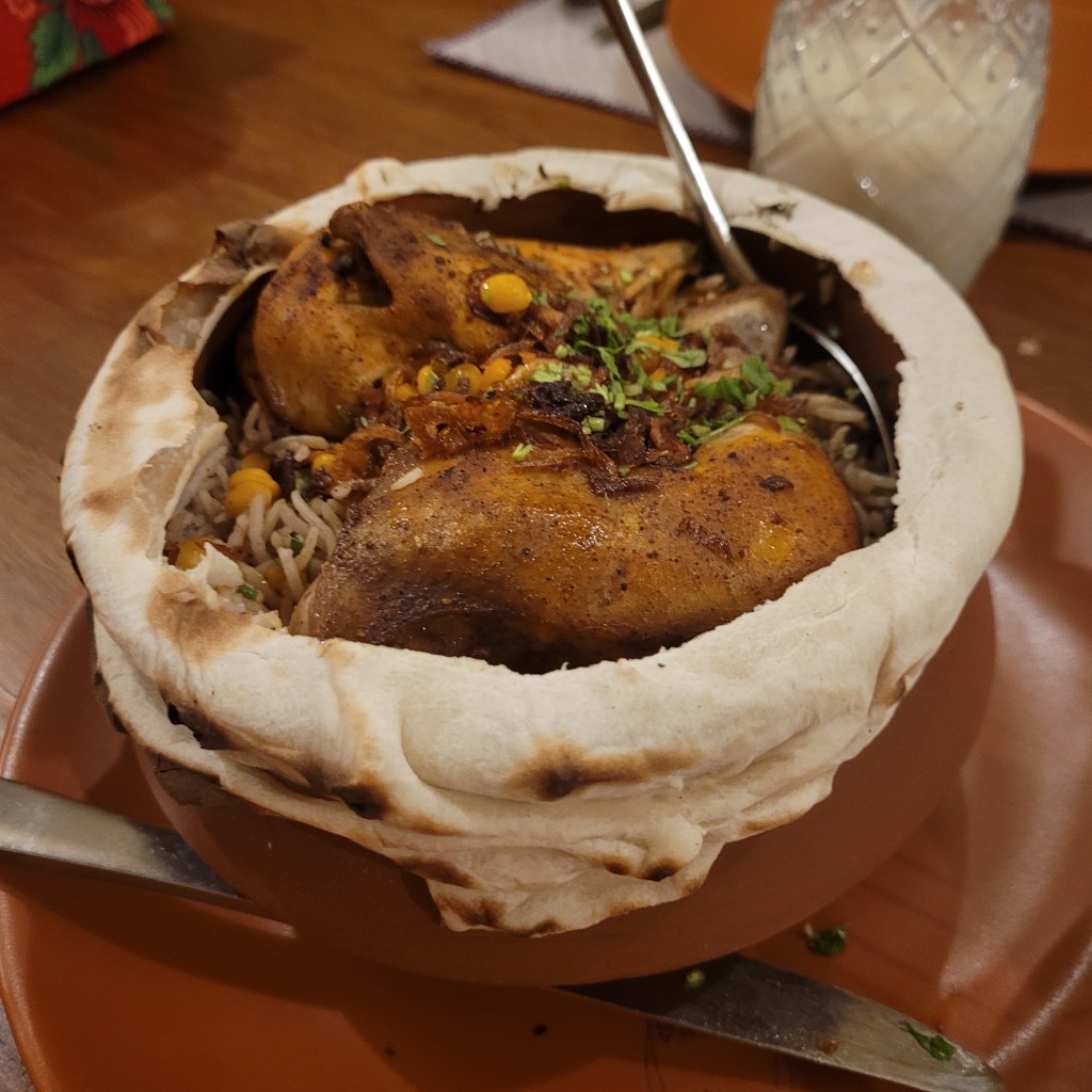 Un plat de poulet servi dans un pot en terre cuite, recouvert de pain plat, avec du riz et des garnitures.