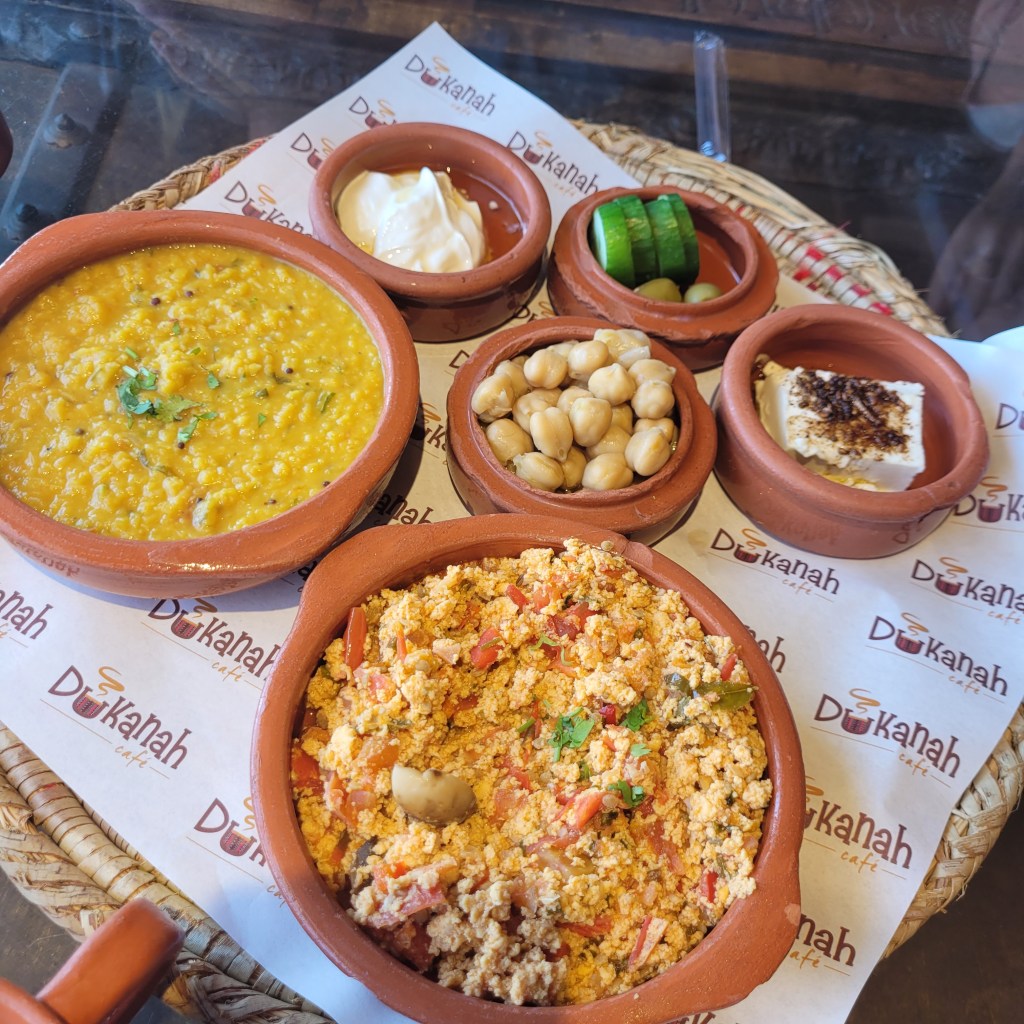Assiette de plats traditionnels dans des bols en terre cuite, comprenant du dal jaune, du bhurji aux tomates, des pois chiches, des tranches de concombre, des olives, une sauce et du yaourt.