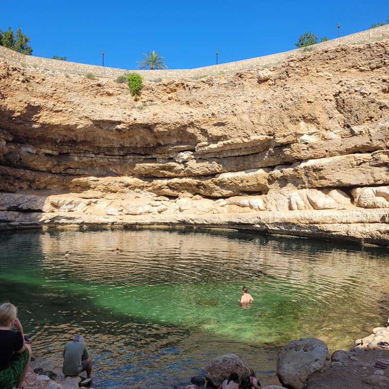 Un paysage de baignade dans une grande grotte avec des parois rocheuses, où plusieurs personnes se baignent dans une eau claire et turquoise, sous un ciel bleu ensoleillé.