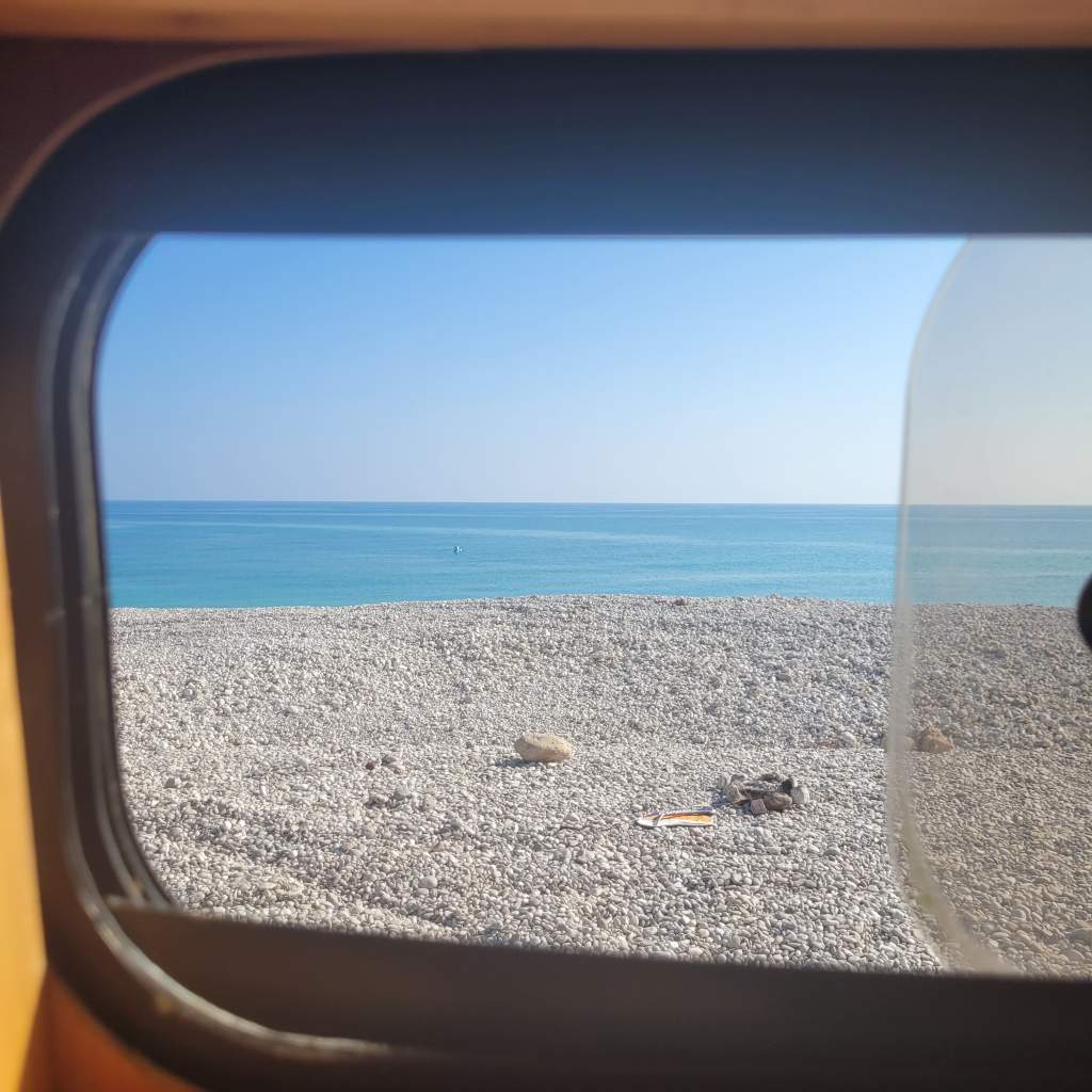 Vue à travers une fenêtre sur une plage de galets et une mer turquoise, sous un ciel clair.