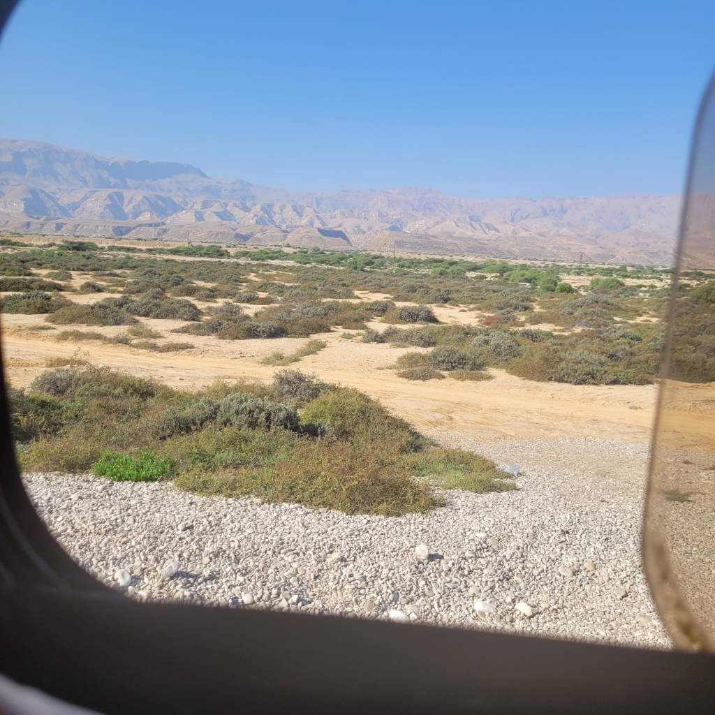 Vue du paysage désertique à travers une fenêtre d'avion, avec des collines en arrière-plan sous un ciel bleu.