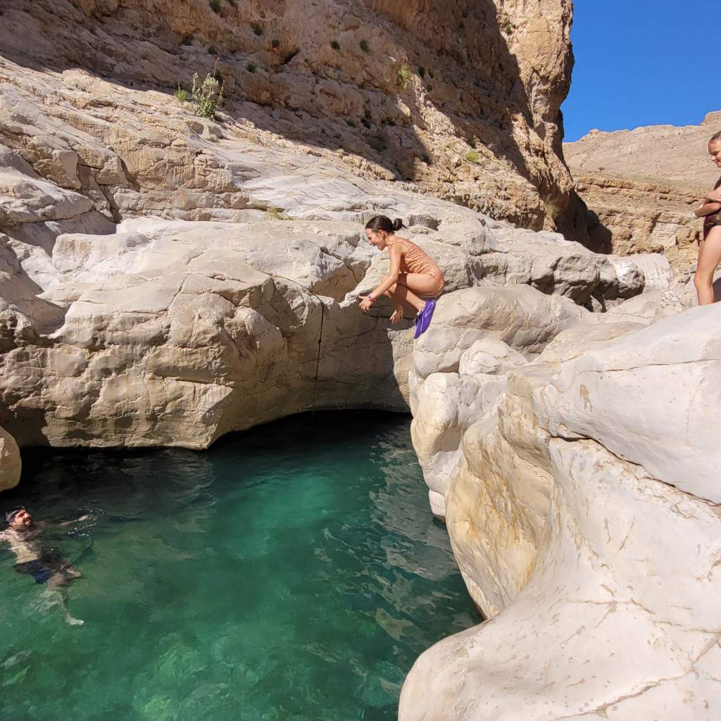 Une jeune fille sautant d'un rocher dans une piscine naturelle turquoise, avec des parois rocheuses et un autre nageur visible à proximité.