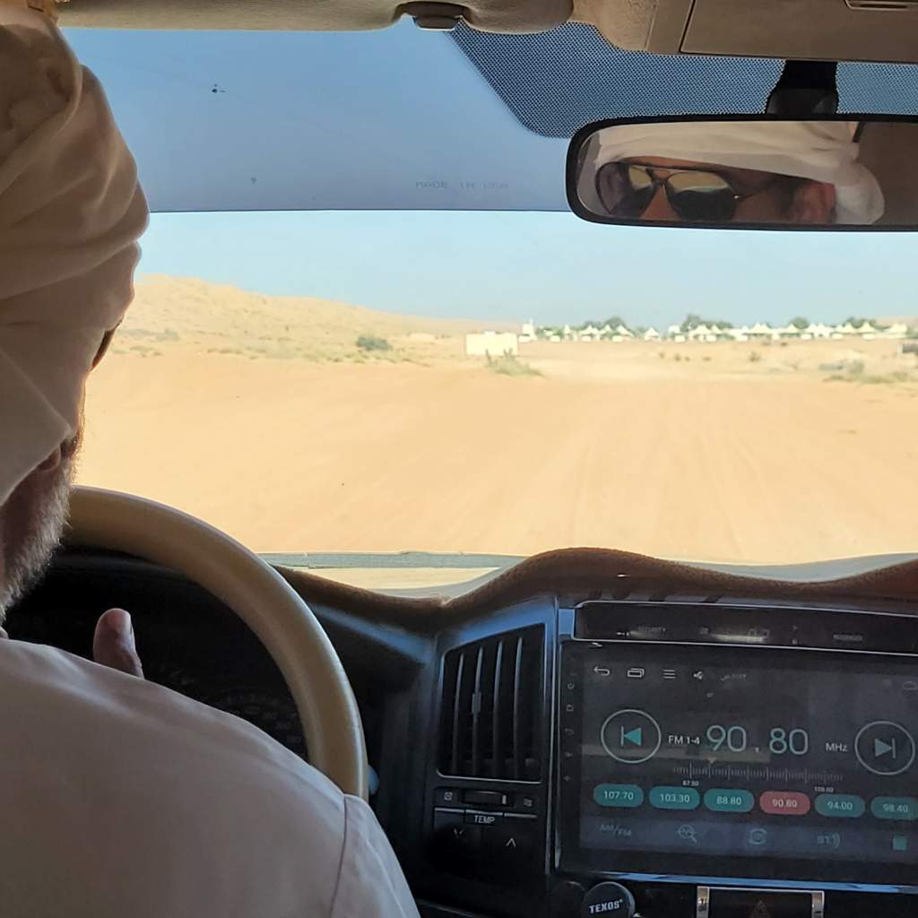 Vue de l'intérieur d'un véhicule, le conducteur portant un turban, dirigeant sur une route de sable avec un tableau de bord visible.