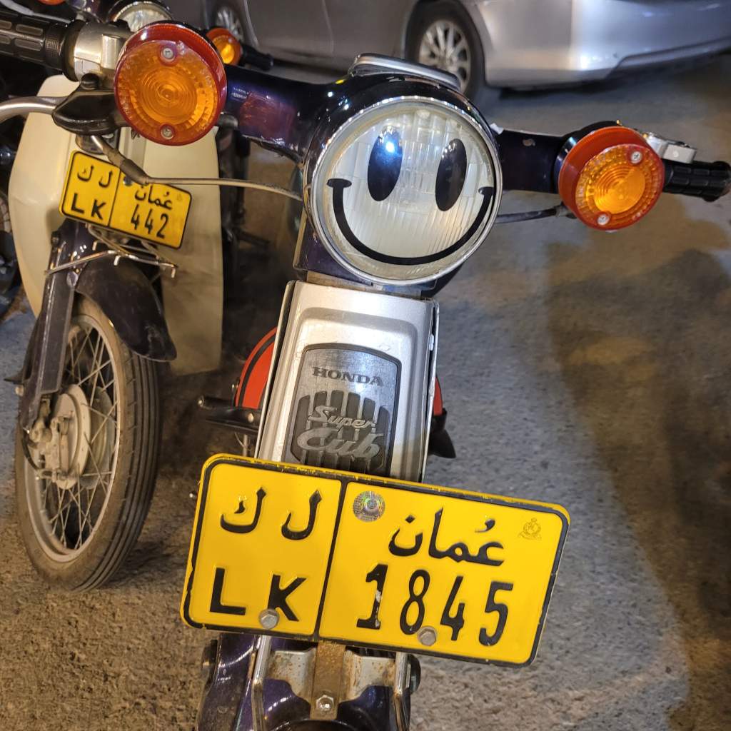 Un scooter Honda avec un phare en forme de smiley, garé dans une rue. Les plaques d'immatriculation affichent des chiffres et des lettres en arabe et en anglais.