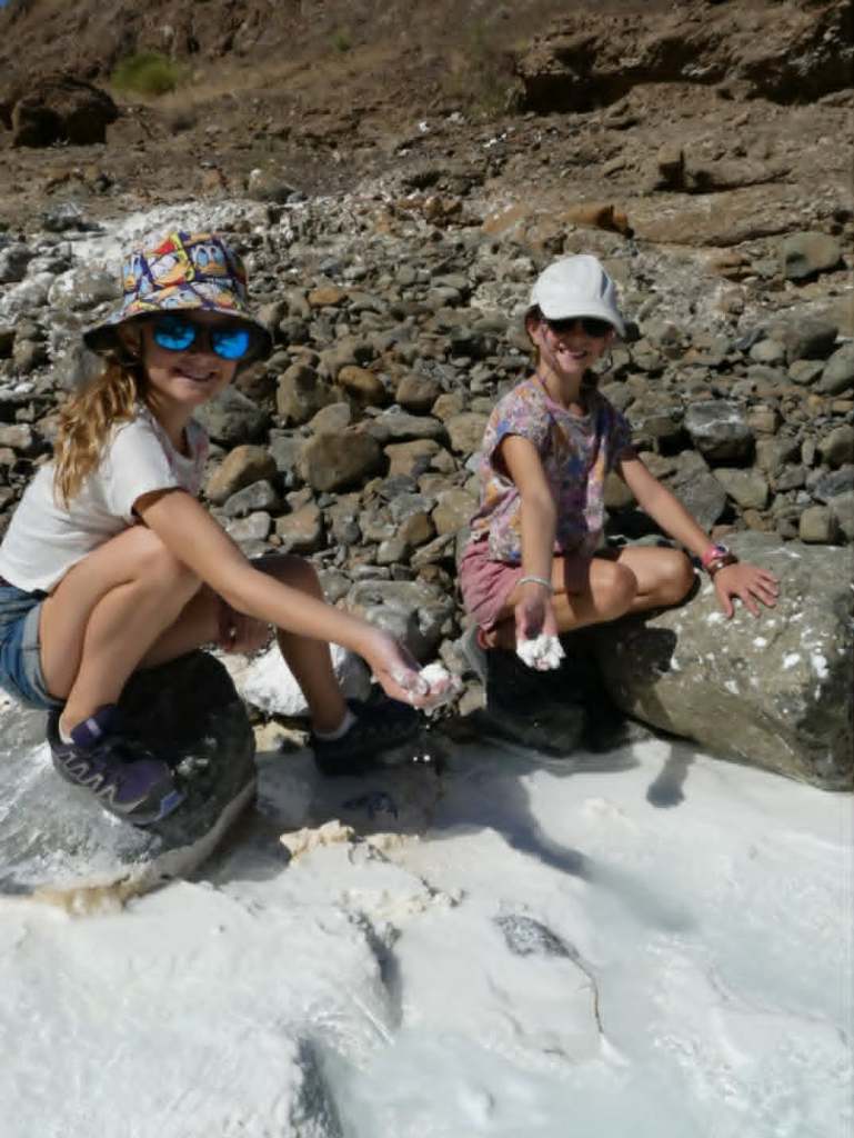 Deux jeunes filles souriantes assises sur des rochers, tenant des morceaux de matière blanche, avec un paysage de pierres et de terrain sec en arrière-plan.