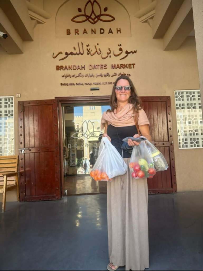 Femme se tenant devant le marché de dattes Brandah, portant des sacs en plastique avec des fruits à l'intérieur.