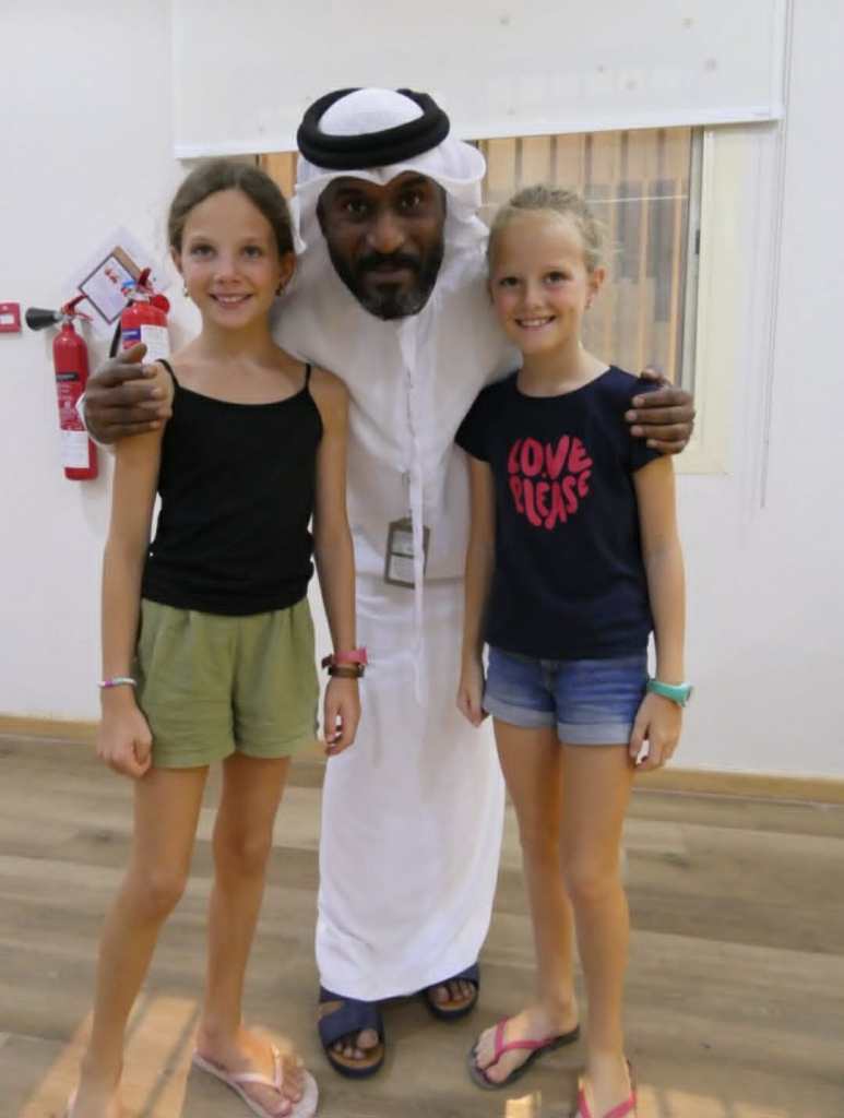 Deux filles souriantes posent avec un homme en tenue traditionnelle, dans un intérieur lumineux.