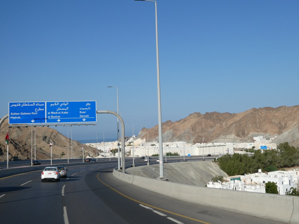 Panneaux de signalisation routière indiquant les directions vers le port Sultan Qaboos, Matrah, et d'autres destinations, avec des montagnes en arrière-plan.