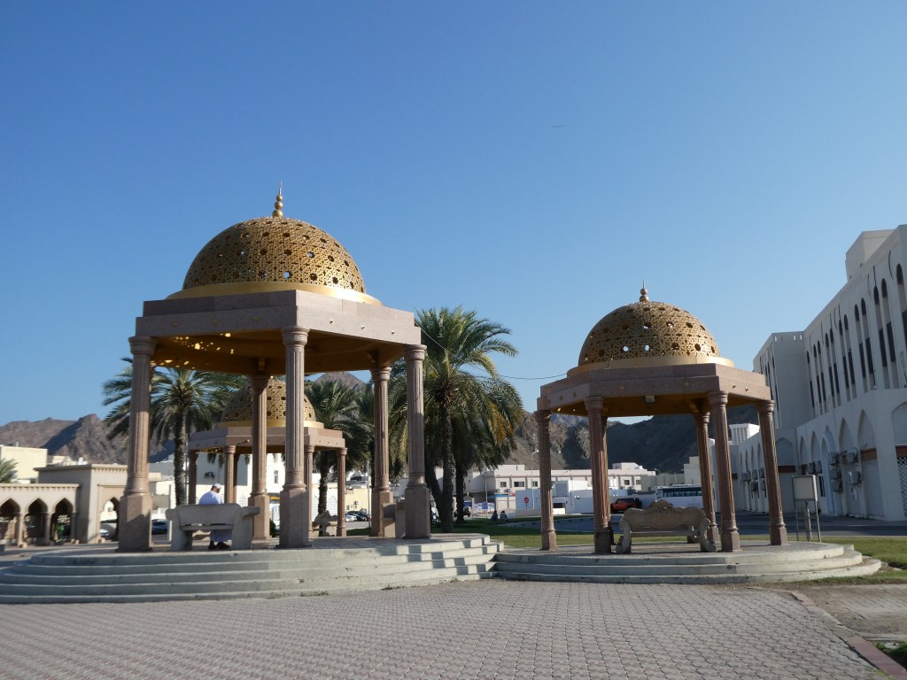 Deux kiosques avec des dômes dorés dans un espace ouvert, entourés de palmiers et de bâtiments, sous un ciel bleu.