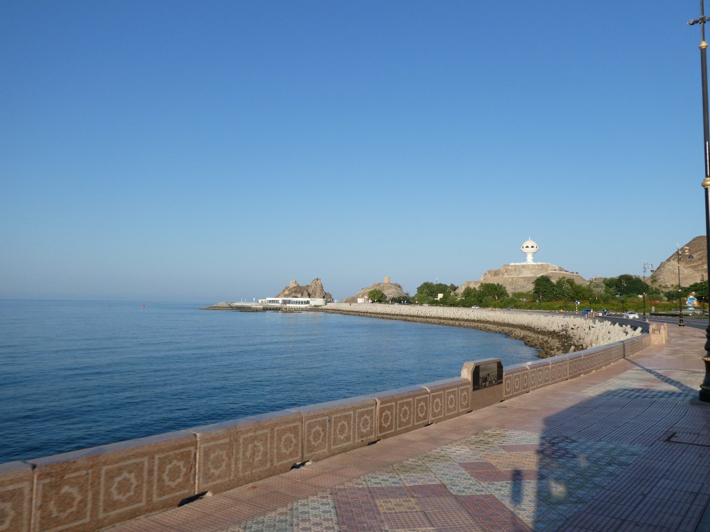 Vue panoramique d'une promenade au bord de la mer avec des collines et un ciel clair.