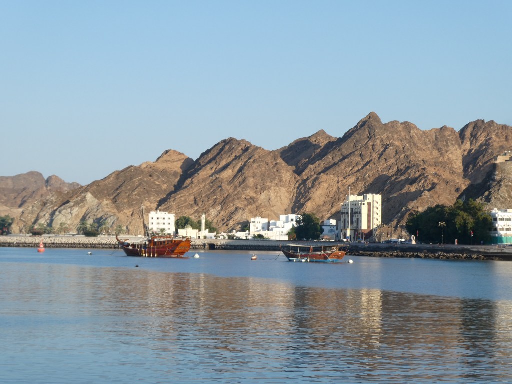 Vue du port avec des dhows traditionnels sur l'eau et des montagnes en arrière-plan sous un ciel dégagé.