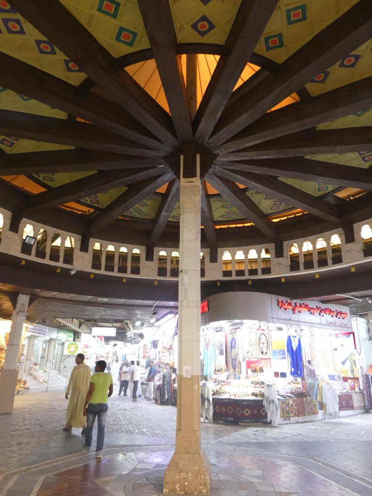 Vue intérieure d'un souk avec un plafond en bois en forme de dôme et des colonnes, entouré de boutiques lumineuses.