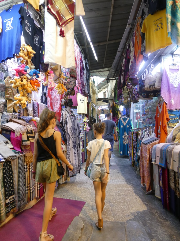 Deux jeunes filles marchant dans un marché animé, entourées de vêtements colorés et d'articles artisanaux accrochés le long des murs.