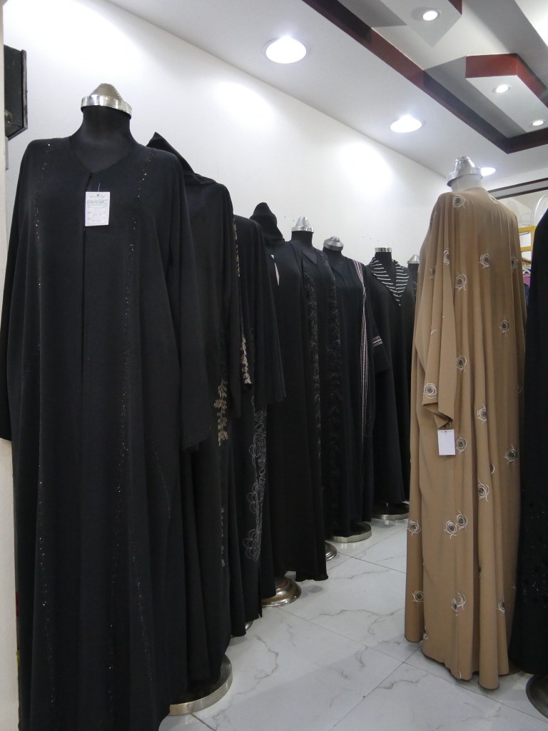 Une rangée de vêtements traditionnels exposés dans un magasin, avec plusieurs robes noires et une robe beige ornée, sur des mannequins.