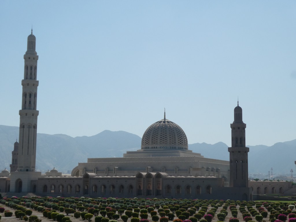 Vue d'une mosquée avec un dôme et des minarets, entourée de jardins et de montagnes en arrière-plan.