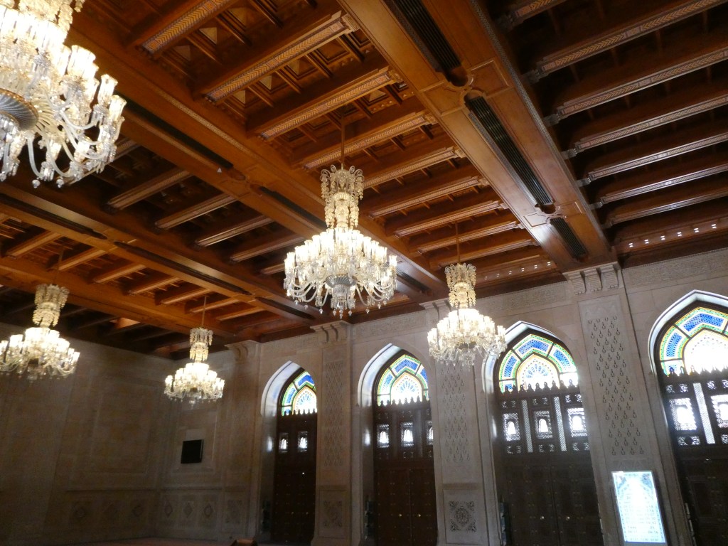 Intérieur d'une salle avec un plafond en bois décoré et des lustres en cristal, illuminant de grandes fenêtres ornées de motifs colorés.
