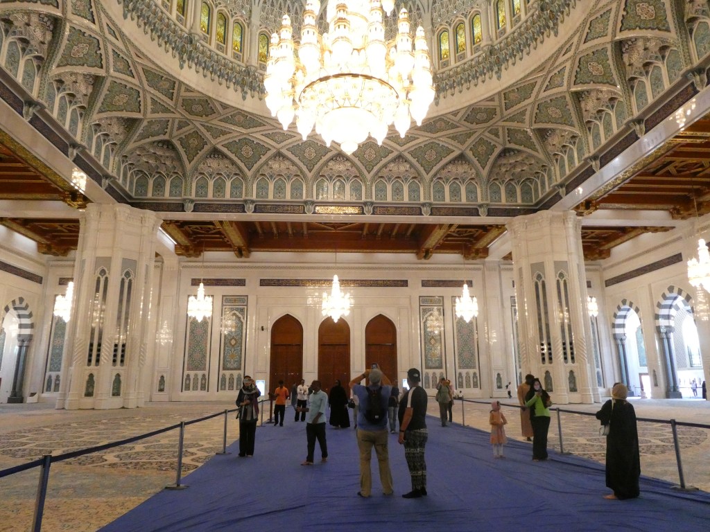 Intérieur d'une mosquée avec un plafond orné de détails complexes et un grand lustre, où des visiteurs explorent l'espace.
