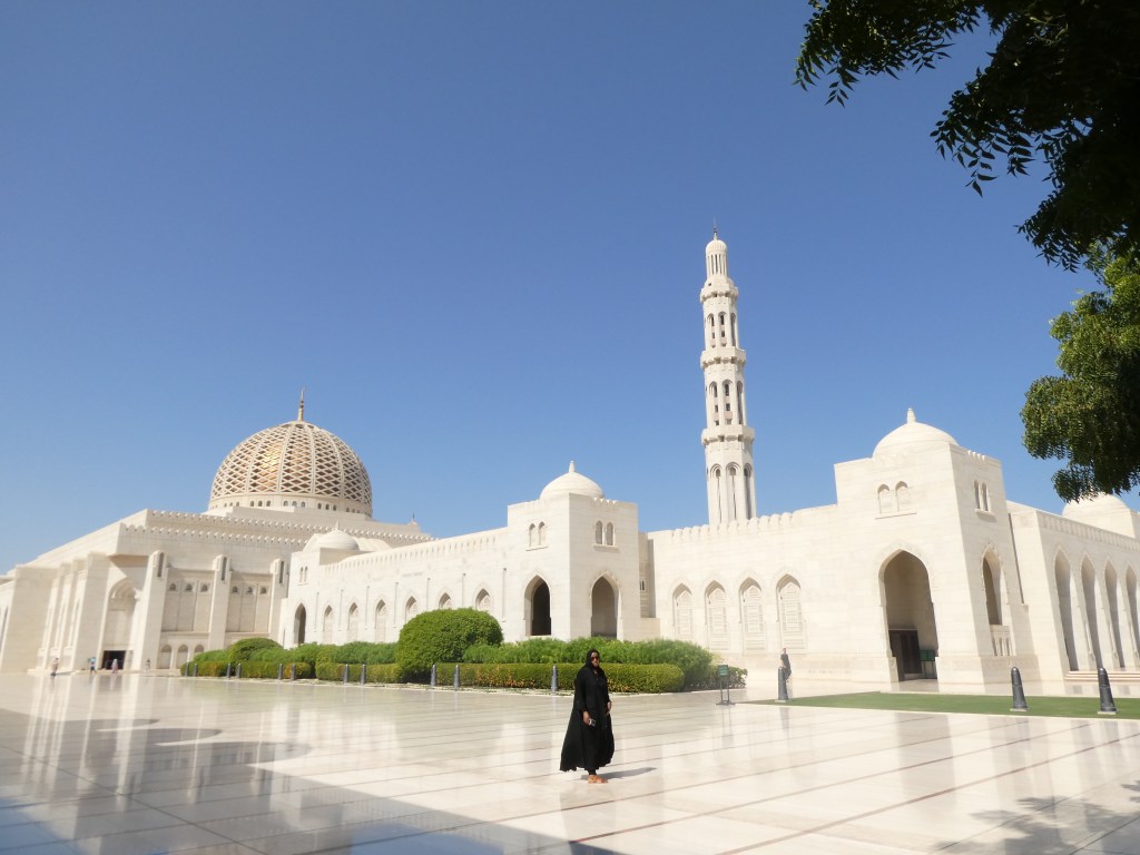 Vue de la Grande Mosquée du Sultan Qabous avec un dôme distinctif et un minaret, une femme en habit traditionnel marchant sur une cour en marbre.