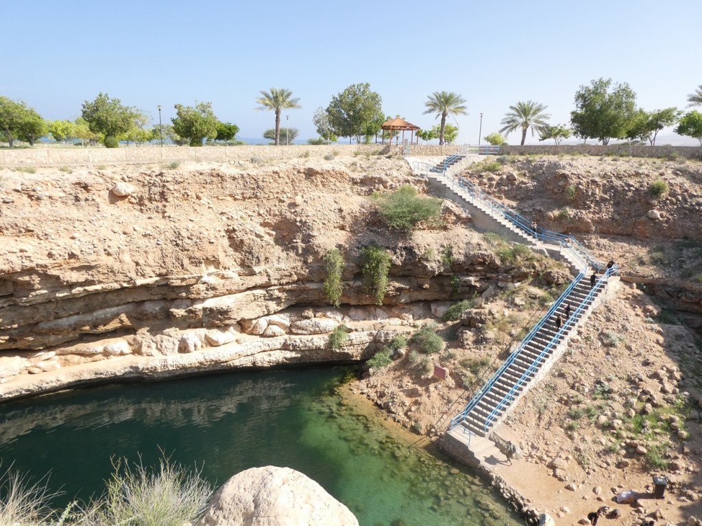 Vue d'une piscine naturelle entourée de rochers et de végétation, avec des escaliers menant au bord de l'eau et un pavillon en arrière-plan.