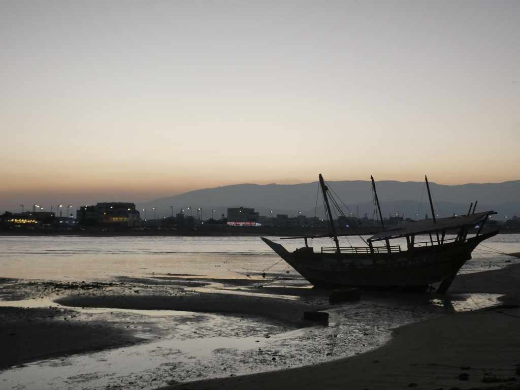 Un bateau traditionnel échoué sur la plage au coucher du soleil, avec des montagnes et des lumières de la ville en arrière-plan.