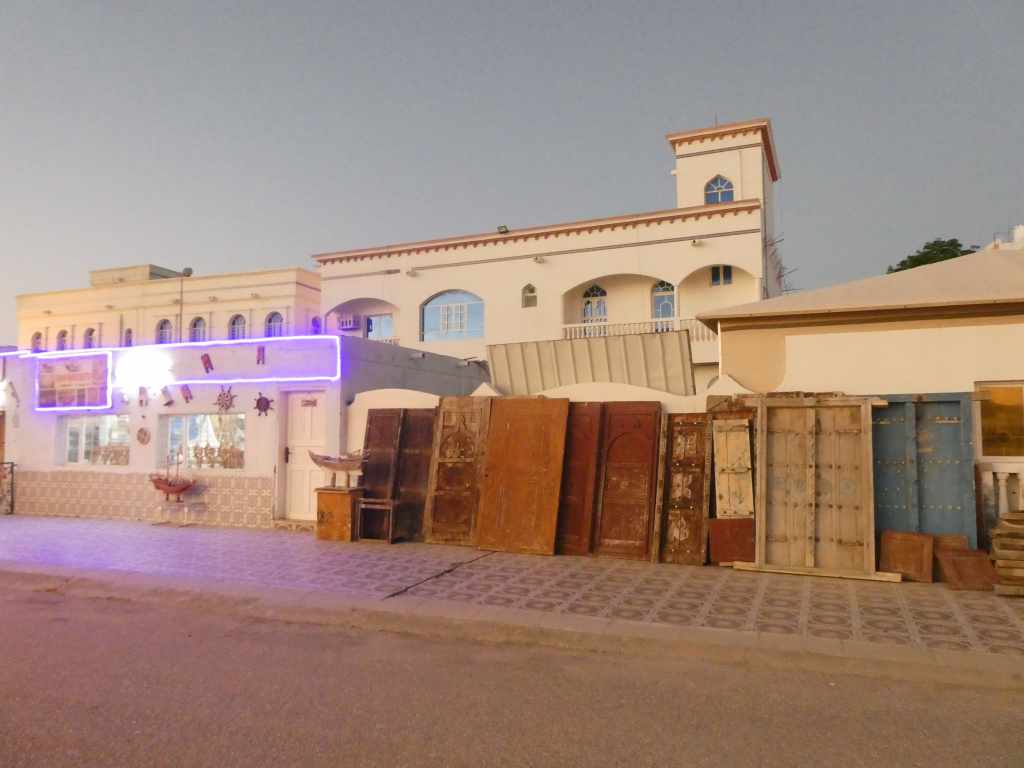 Une rue avec des bâtiments aux façades blanches, décorées de lumières. Plusieurs portes en bois de différentes couleurs sont alignées devant, tandis qu'un bâtiment à deux étages est visible en arrière-plan.