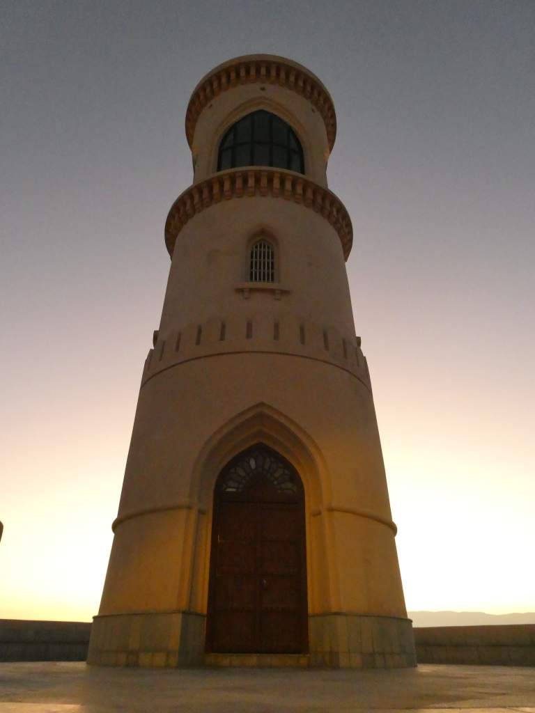 Un phare en pierre, de forme cylindrique, avec une porte en bois et une grande fenêtre, éclairé par la lumière dorée du coucher de soleil.