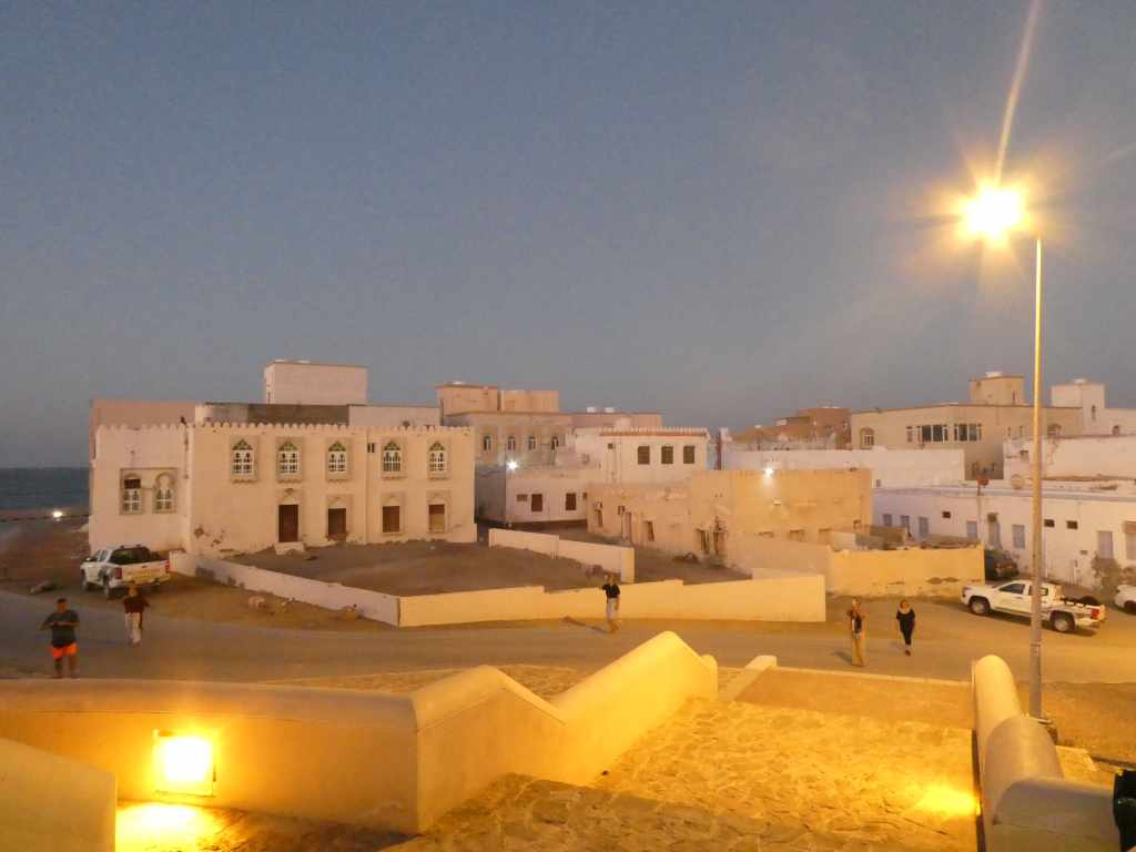 Vue d'une ville côtière au crépuscule, avec des bâtiments traditionnels blanches et des personnes se promenant dans la rue.