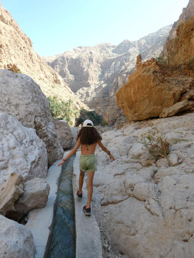 Une jeune fille marche sur un sentier étroit au milieu des montagnes rocailleuses, avec un canal d'eau à sa gauche.