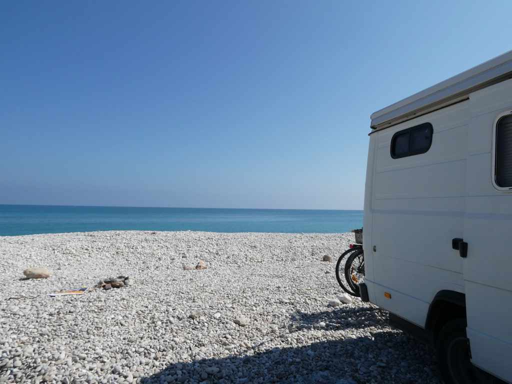 Un fourgon blanc garé sur une plage de galets, avec une vue sur la mer bleue et le ciel dégagé.