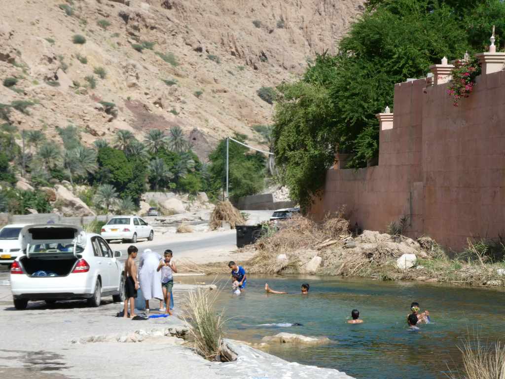 Enfants jouant dans une rivière près d'une voiture garée sur une route, avec des montagnes en arrière-plan.