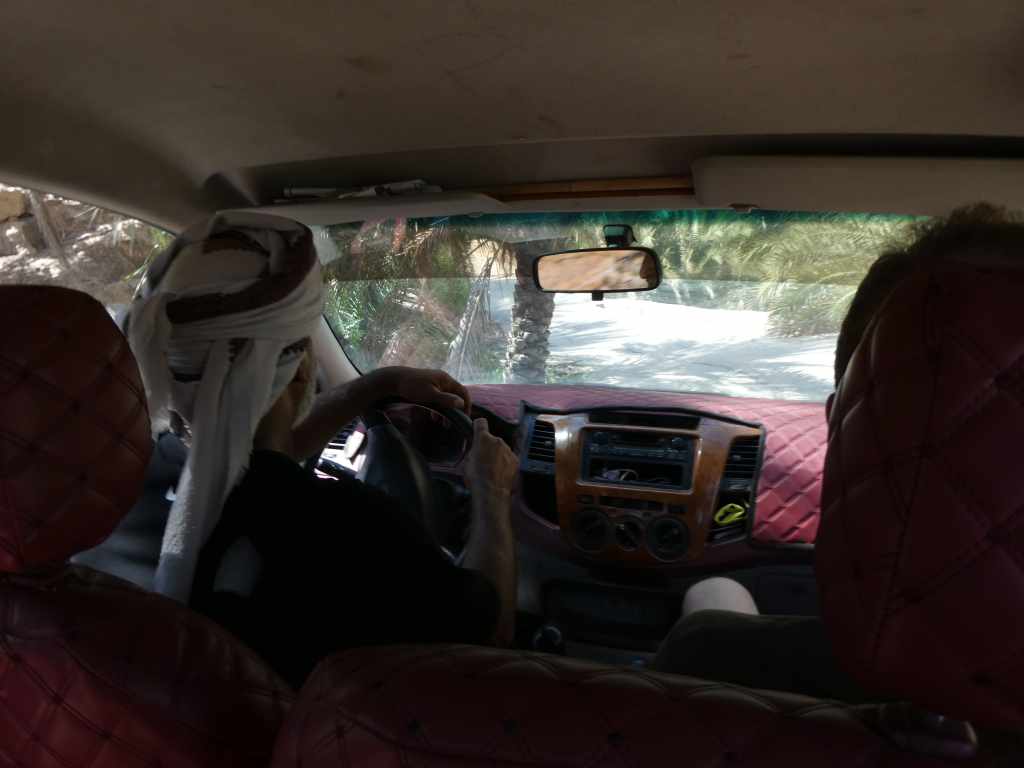 Vue intérieure d'une voiture avec un conducteur vêtu d'une robe traditionnelle et un passager à l'arrière, montrant un chemin bordé de palmiers.