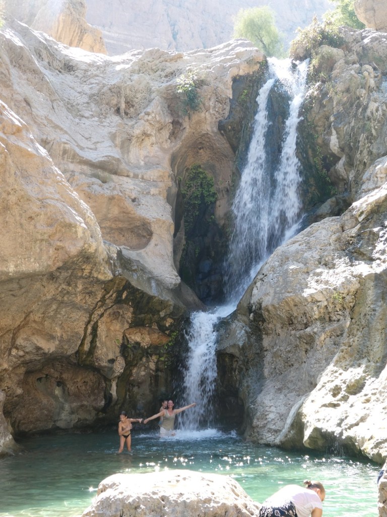 Une cascade se déverse dans une piscine naturelle où deux personnes s'amusent, entourées de rochers et de verdure.